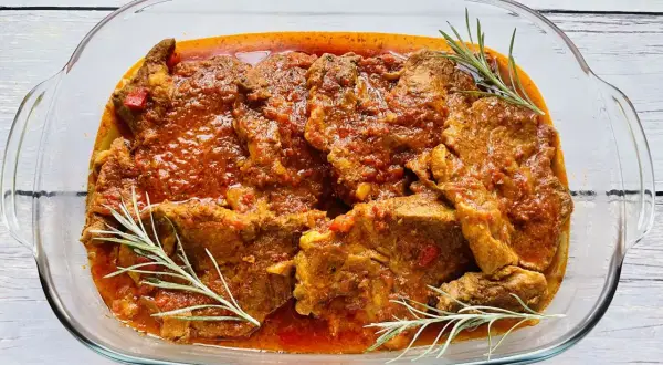 Karkówka w sosie pomidorowym z cebulą, duszona do miękkości, podana w szklanej brytfannie z gałązkami rozmarynu.