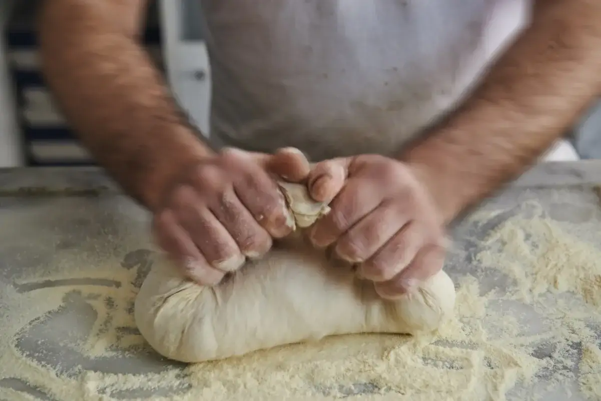 Dłonie ugniatają ciasto na spod od pizzy, rozsypując mąkę na blacie.