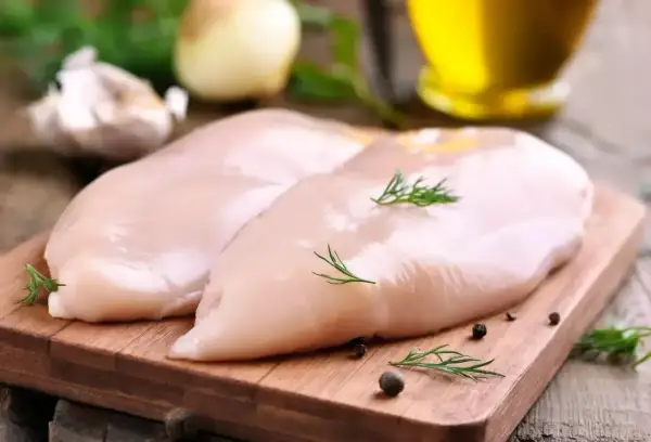 Dwa surowe filety z kurczaka, bogate w białko, leżą na drewnianej desce. Obok czosnek, cebula i oliwa.